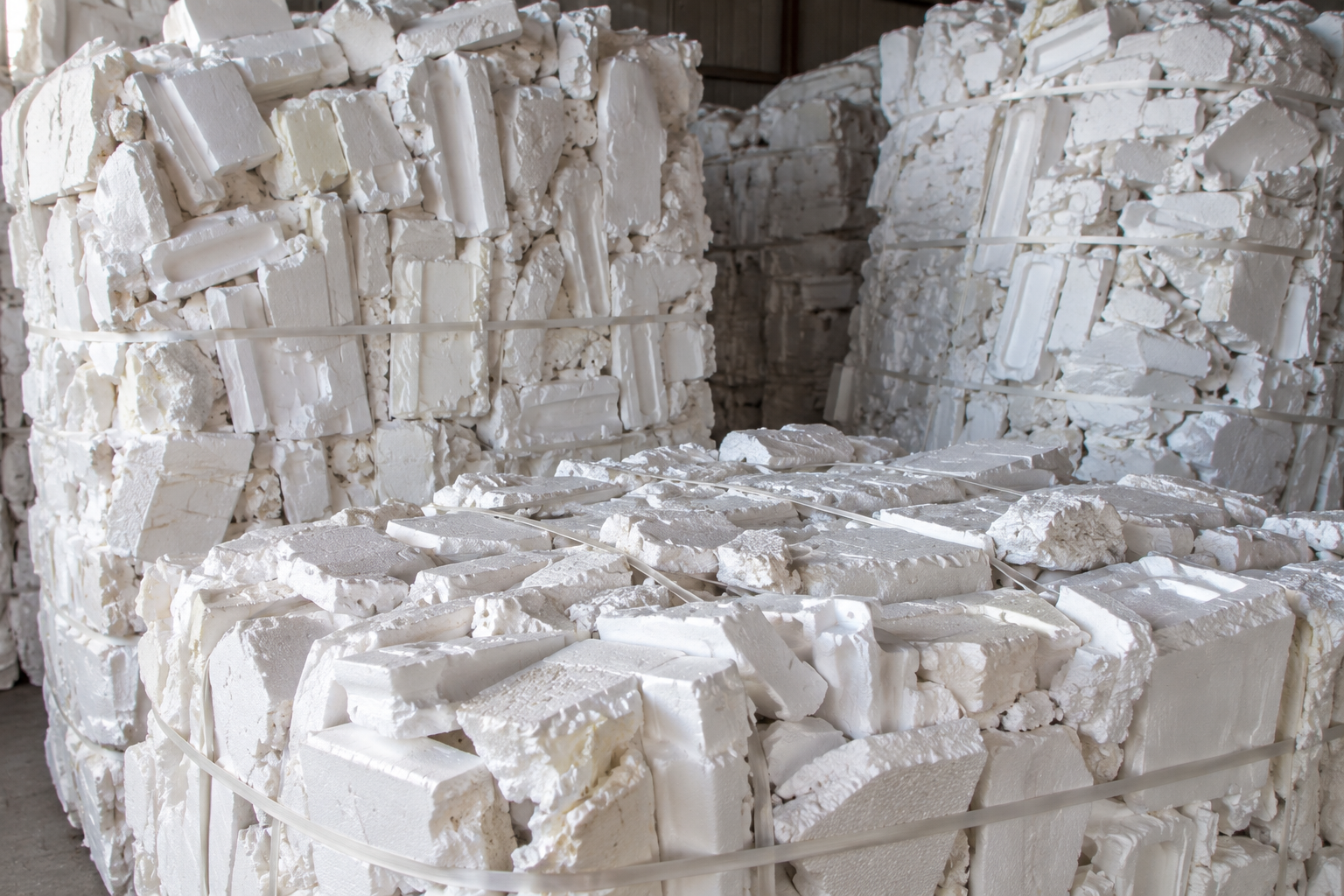 Large stacks of compacted white polystyrene foam blocks are piled together, creating a textured and irregular surface in an industrial styrofoam recycling facility. These blocks have been processed using high-quality densifiers, such as those from Better Densifiers, to ensure efficient and sustainable recycling.