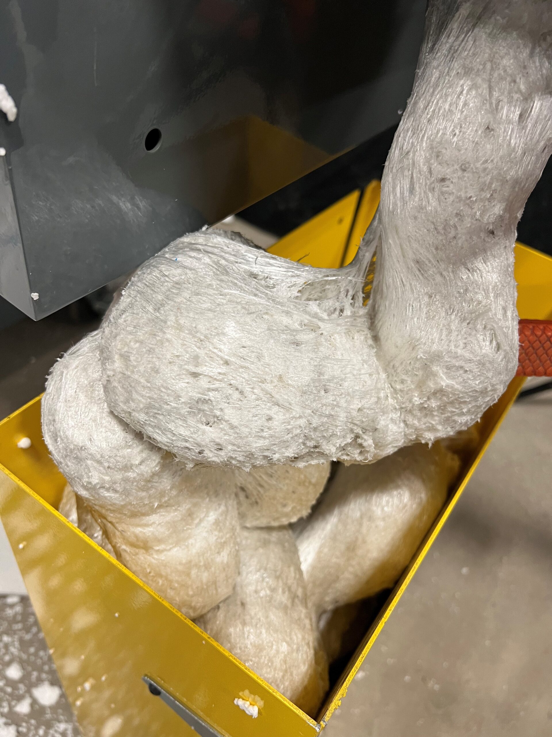 Thick, fibrous, white material overflows from a gray PolyWiz machine into a yellow bin, with strands and clumps of styrofoam piled up inside the container as part of the styrofoam recycling process.