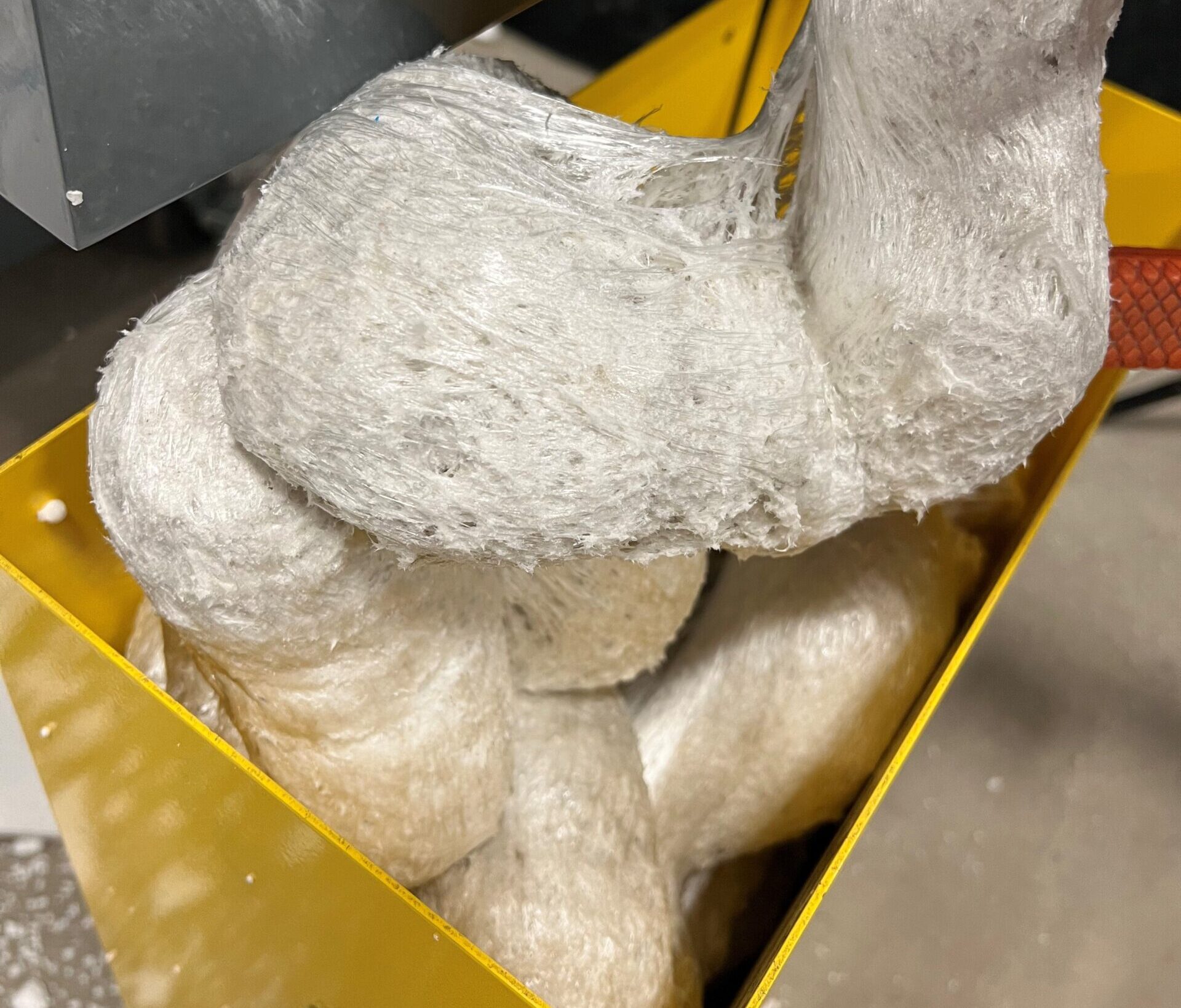 White, fibrous material tightly packed inside a yellow metal container, possibly industrial fibers or insulation. The texture appears soft and fluffy, with reflective surfaces under bright lighting—reminiscent of processed styrofoam often found in styrofoam recycling or used with Better Densifiers for efficient waste management.