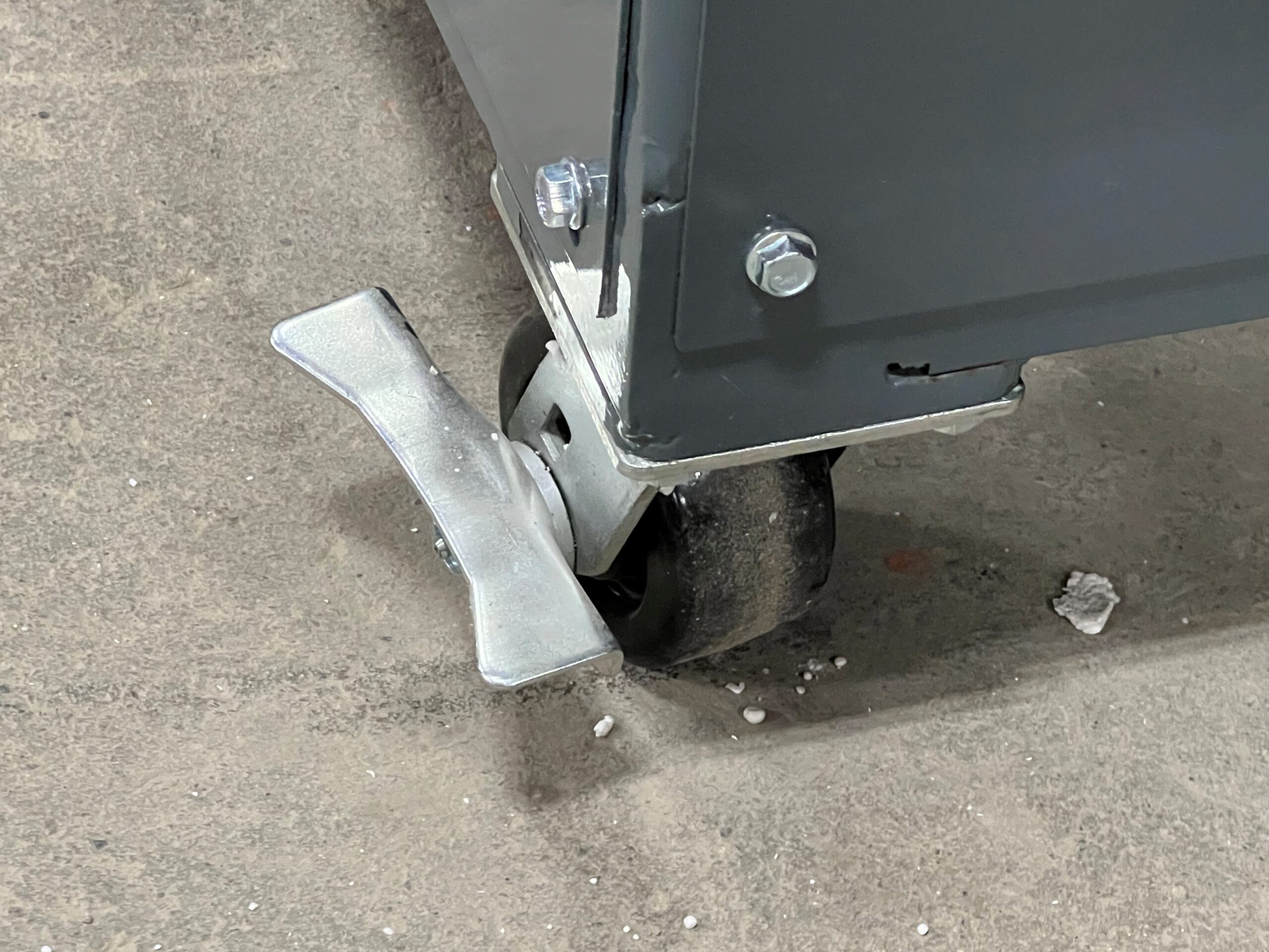 A close-up of a caster wheel with a metal brake lever attached to the bottom of a gray metal cart, on a rough concrete floor—ideal for maneuvering equipment such as Better Densifiers used in styrofoam recycling.