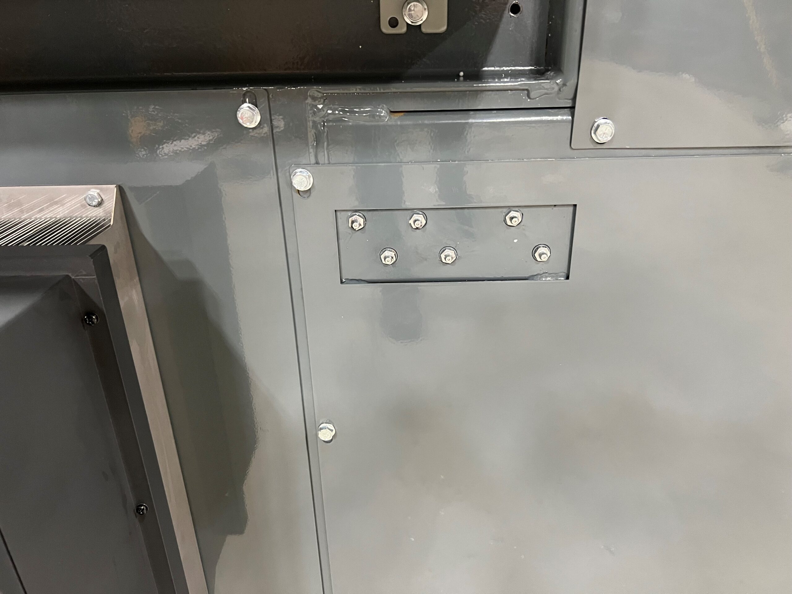 A close-up of a gray metal surface with several bolts and panels, including a rectangular panel fastened with multiple screws. The image shows part of a machine or industrial equipment, possibly related to PolyWiz densifiers designed for processing styrofoam.