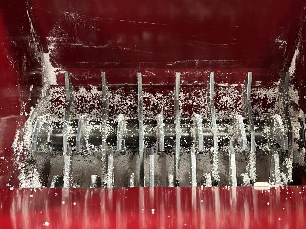 Close-up of a red industrial machine, such as a PolyWiz styrofoam densifier, with metal blades and salt-like particles scattered inside. The blades are arranged vertically and horizontally for efficient grinding or shredding.