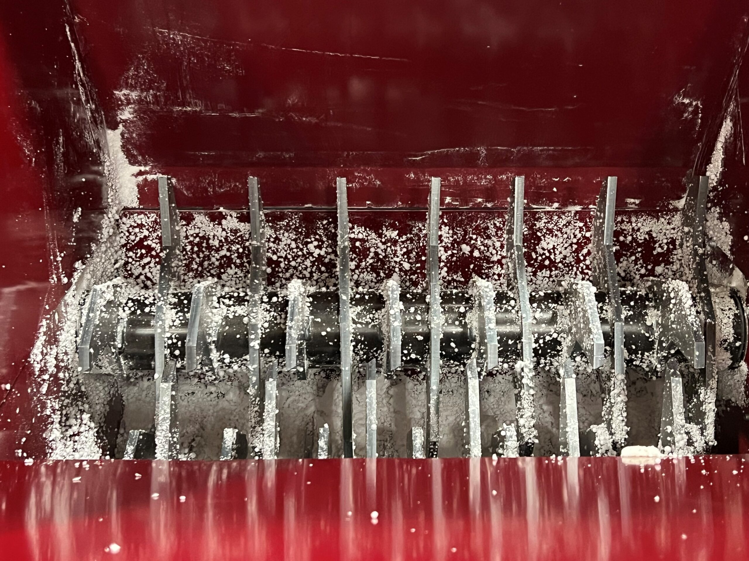 Close-up of a red industrial machine, such as a PolyWiz styrofoam densifier, with metal blades and salt-like particles scattered inside. The blades are arranged vertically and horizontally for efficient grinding or shredding.