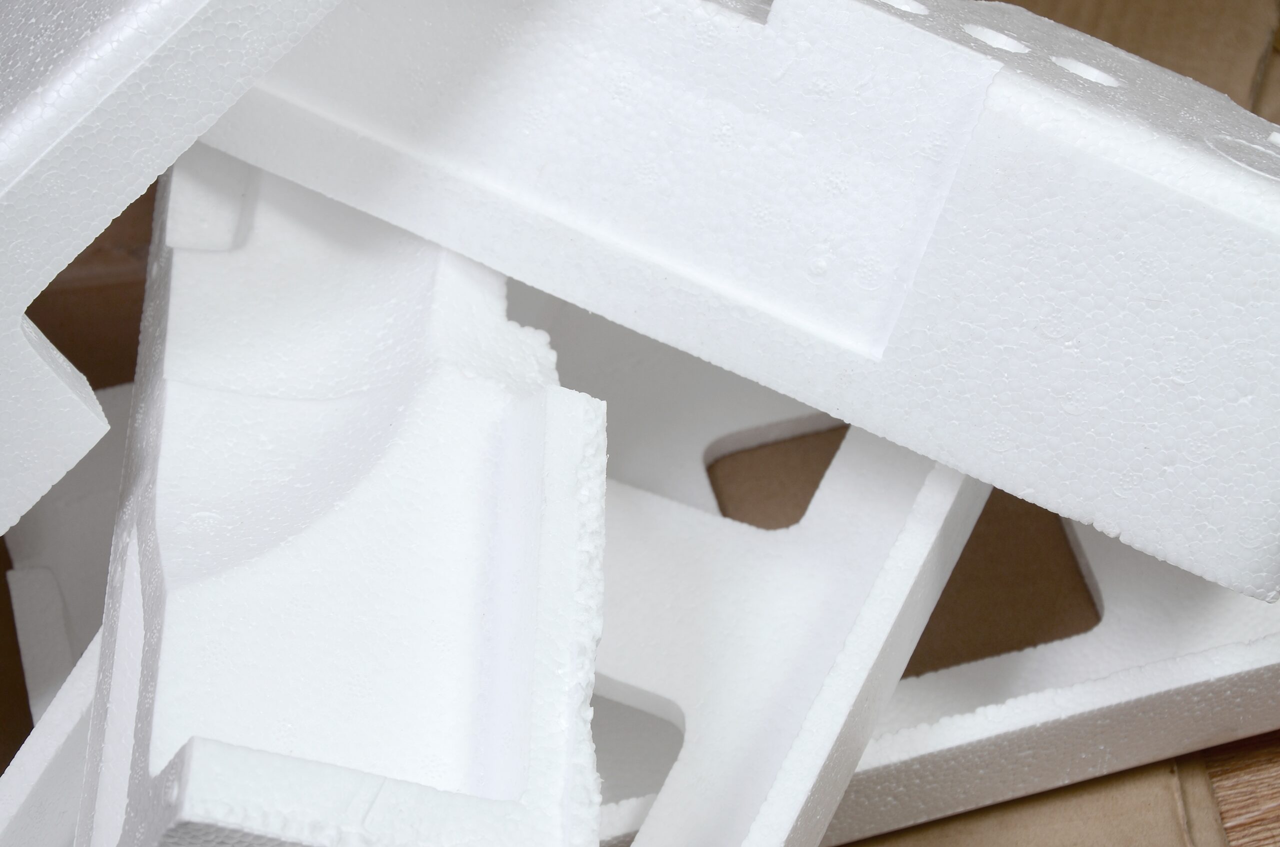 Close-up of several white polystyrene foam packaging pieces, sometimes referred to as styrofoam, stacked on top of each other, with some cardboard visible in the background.