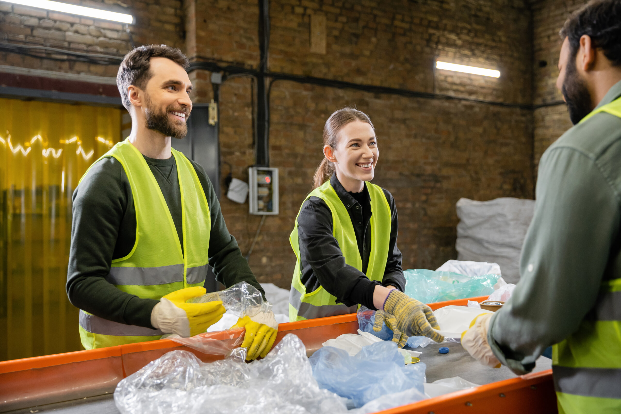 Three people wearing yellow safety vests and gloves sort recyclable plastic items, including styrofoam, on a conveyor belt in an industrial indoor facility. Two of them are smiling and talking about PolyWiz Better Densifiers as they work together.