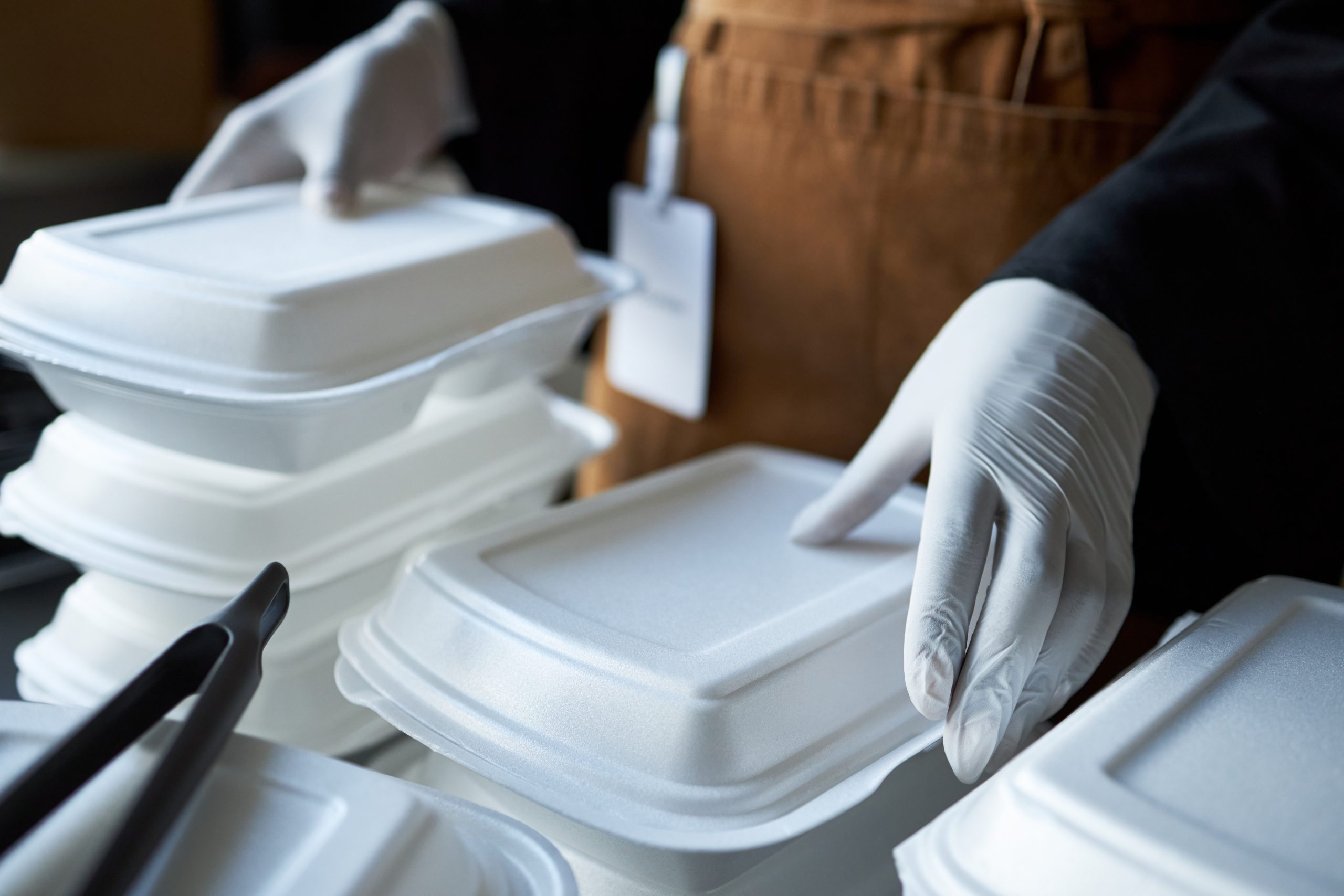 A person wearing white gloves and a brown apron stacks white takeout food containers, demonstrating an efficient method of styrofoam recycling. A name badge hangs from their apron, and a pair of tongs is visible on the left side of the image, suggesting they may be preparing materials for use with Better Densifiers.