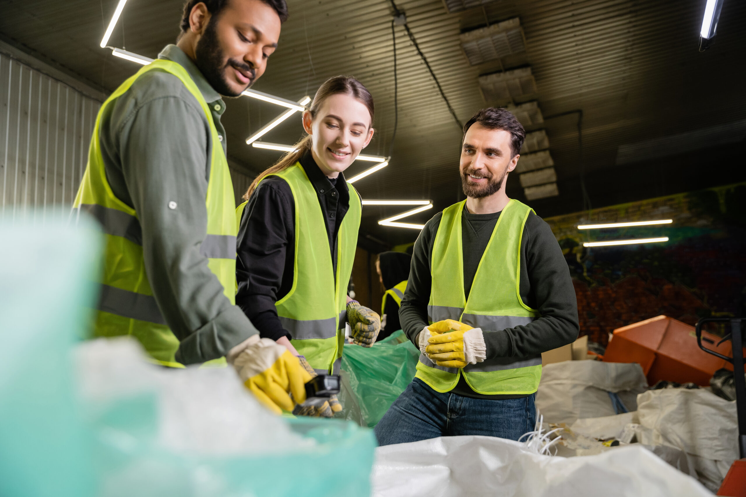 Three people wearing yellow safety vests and gloves sort recyclables, including styrofoam, together in an industrial facility equipped with densifiers, smiling and working as a team under bright overhead lighting.