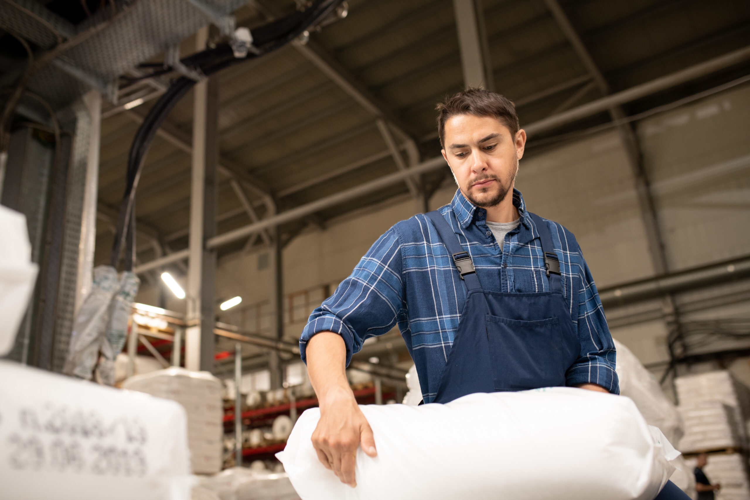 Man in a plaid shirt and blue overalls lifting a large white sack in an industrial warehouse, surrounded by machinery—including Better Densifiers for styrofoam—and shelves stacked with more sacks.