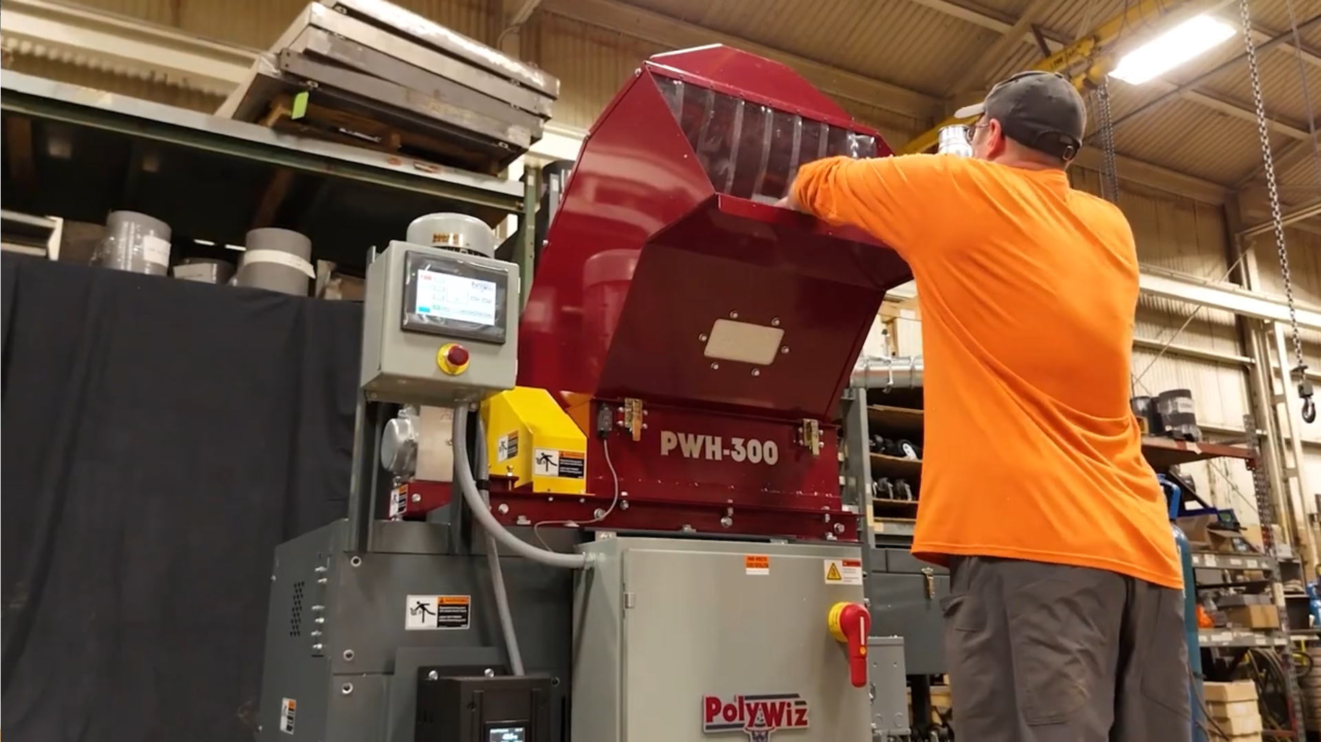 A person wearing an orange shirt operates a large red industrial machine labeled PWH-300 at a factory, reaching into its open top section. This PolyWiz Better Densifiers unit is part of the styrofoam recycling process. Shelves and equipment are visible in the background.