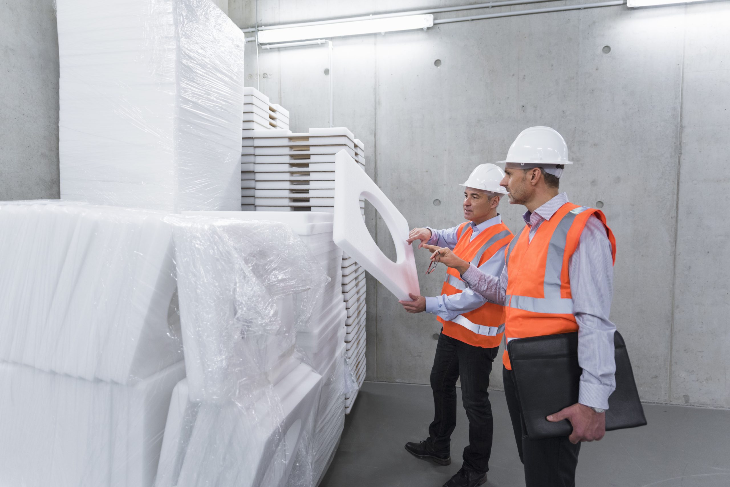 Two workers in safety vests and hard hats inspect PolyWiz foam packaging in an industrial warehouse. One holds a styrofoam piece, while the other observes and carries a black folder. Stacks of foam are visible around them, highlighting the need for densifiers in efficient material handling.