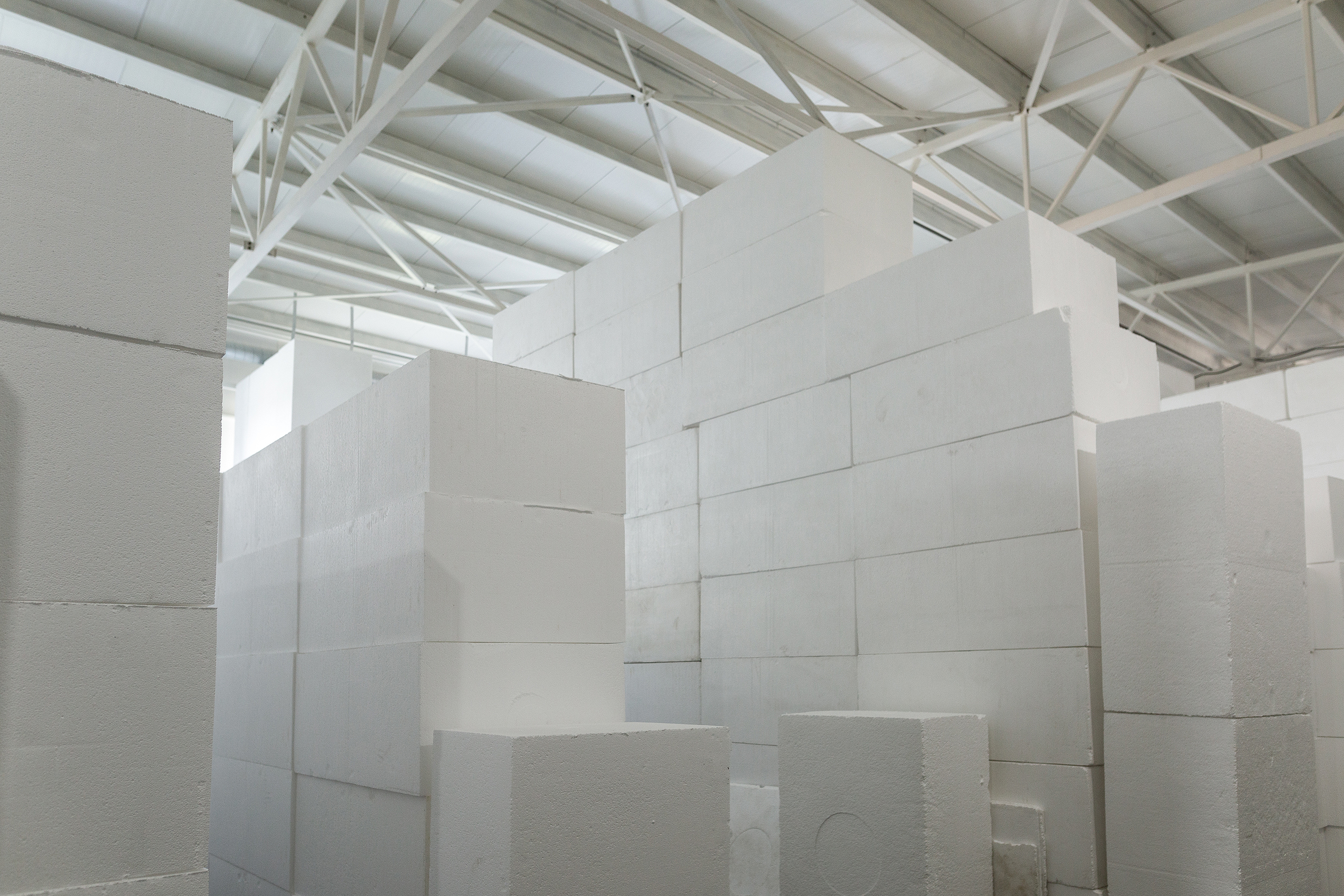 Large white rectangular blocks, created through a PolyWiz densifier as part of a styrofoam recycling process, are stacked in a warehouse with a high ceiling and exposed beams, forming tall, geometric structures.
